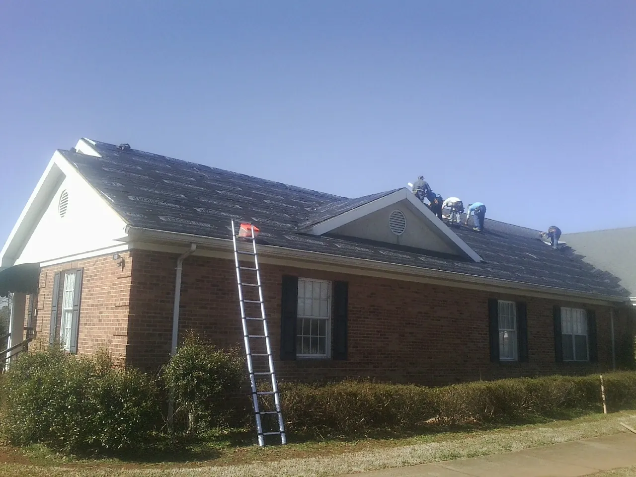 Crew tearing shingles off a brick ranch roof with a ladder set — Emergency Roof Repair work by our crew in Flint